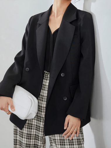 Veste tailleur femme élégante et décontractée sur une femme debout contre un mur gris tenant un sac à main blanc