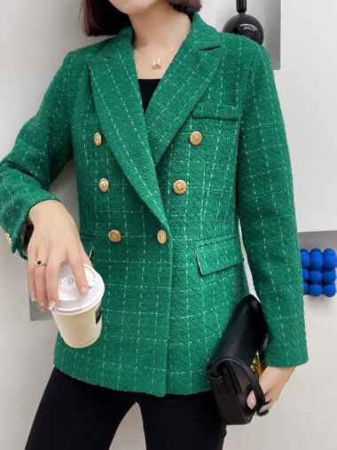 Veste habillée style blazer vert à carreaux portée par une jeune femme brune qui porte son sac dans la main gauche et une tasse de café dans la main droite. Elle est adossée à un mur blanc.
